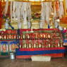 An altar with offering bowls, silk scarves, incense burner, and tormas at the foot of a large statue.