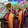 A procession of monks wearing yellow and red hats and carrying cymbals, religious texts, and parasols.